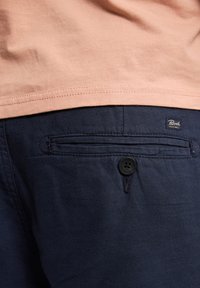 Pantalones azul marino hechos de una tela de textura suave; incluyen un bolsillo trasero con un botón negro y una pequeña etiqueta de marca en la cinturilla.