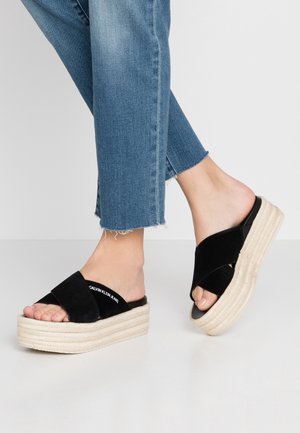 Fødder iført sorte platformsandaler med krydsede remme og hvid sål, kombineret med blå jeans med frynsede kanter mod en ensfarvet baggrund.