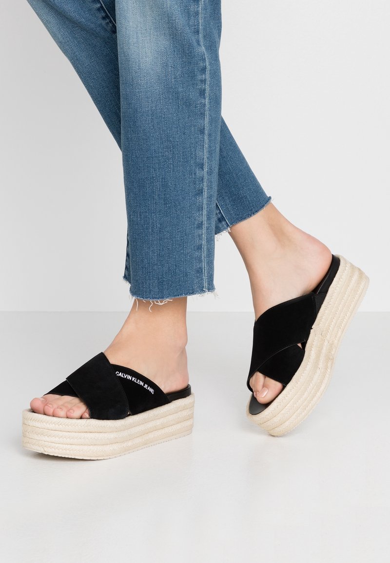 Pieds portant des sandales plateformes noires avec des lanières croisées et une semelle blanche, associées à un jean bleu à ourlet effiloché sur un fond uni.