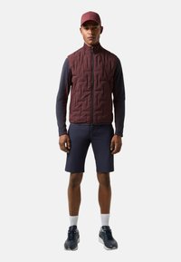 Dunkelroste gesteppte Weste über einem marineblauen Langarmshirt, kombiniert mit marineblauen Shorts und schwarzen Turnschuhen. Die Weste hat einen hohen Kragen und einen Reißverschluss.