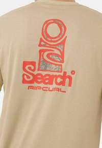 Camiseta de algodón beige con un gran estampado gráfico rojo en la parte posterior, que incluye "Search" y "Ripcurl," con un patrón texturizado en el logotipo.