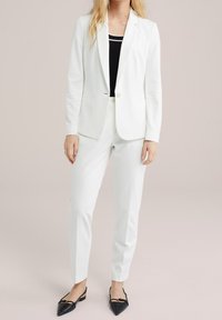 Bílý blejzr a odpovídající kalhoty, s přiléhavým černým topem pod tím. Blejzr má klopové límce a dvě přední kapsy. Černé baleríny.