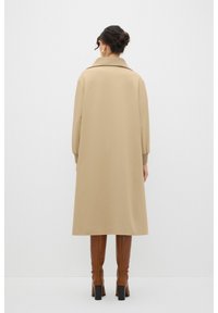 Beige halflange jas met een kraag, lange mouwen en zijzakken. Gedragen boven bruine kniehoge laarzen met blokhak. Glad textuur.
