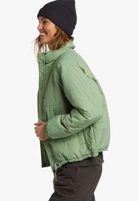 Giacca a vento verde chiaro con colletto alto, polsini elasticizzati e vestibilità rilassata. Presenta una chiusura con zip e dettagli di cucitura visibili.