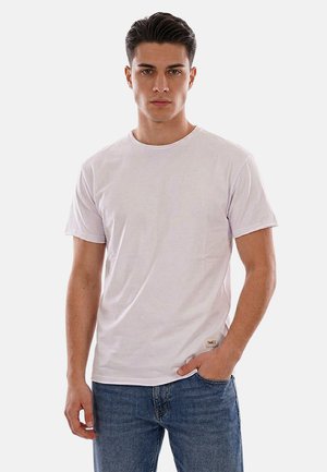 Giovane uomo che indossa una t-shirt bianca semplice e jeans blu, in piedi con una mano in tasca contro uno sfondo chiaro uniforme.