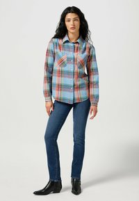 Långärmad rutig skjorta i blått, rött och grönt med två bröstfickor, knäppning med knappar och rund krage, i kombination med smala blå jeans och svarta stövlar.