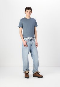 Blå kortermet t-skjorte i bomull, lyseblå baggy jeans og brune snørebåndsstøvler. Antrekket har en avslappet passform og uformell stil.