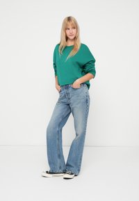 Zelený voľný sveter s dlhými rukávmi, doplnený svetlomodrými rozšírenými džínami a čiernymi plátenými teniskami. Jednoduché pozadie.