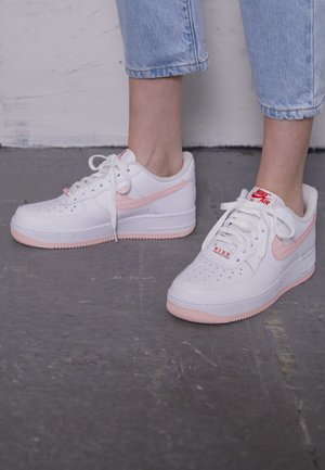 Białe buty Nike Air z jasnoróżowymi podeszwami i znakami, noszone z krótkimi jasnoniebieskimi dżinsami na betonowej podłodze.
