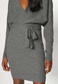 Robe en tricot côtelé gris avec un décolleté en V, des manches longues et un détail de taille à nouer. Le tissu a une texture douce et un design ajusté.