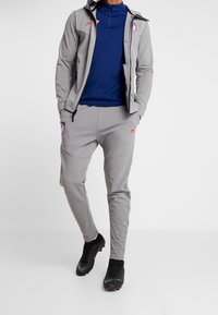 Chándal deportivo gris con chaqueta de cremallera y pantalones a juego, con una parte superior azul y acentos rojos de Nike. Botas negras completan el conjunto.