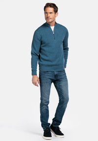 Blauwe ritstrui gemaakt van gebreide stof, met een hoge kraag, ribgebreide manchetten en zoom. Gecombineerd met donkerdenim jeans en zwarte schoenen.