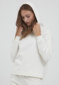 Felpa bianca con cappuccio, dal taglio morbido e rilassato, con polsini a costine e una texture soffice. Il modello tiene il cappuccio, rivelando un design semplice.