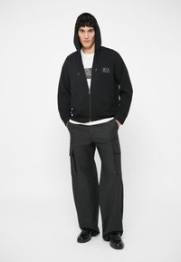 Crna zip-up dukserica s kapuljačom i prednjim džepom; detalj s logotipom na prsima. Uparen s širokim crnim cargo hlačama i crnim cipelama.