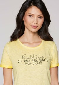 Gelbes Kurzarm-T-Shirt mit rundem Halsausschnitt und umgeschlagenen Ärmeln. Mit einem bedruckten schwarzen Design, das "Dächer auf der ganzen Welt" liest.