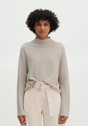 Vrouw met krullend haar, gekleed in een beige coltrui en lichtbeige ceintuurbroek tegen een effen witte achtergrond.
