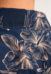 Tessuto navy con grandi motivi floreali beige; cintura elastica con targhetta metallica visibile; texture liscia.