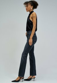 Mujer de perfil, vestida con camiseta negra sin mangas, jeans acampanados gris oscuro, tacones negros puntiagudos y pendientes plateados, sobre fondo gris.