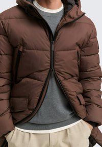 Braune Pufferjacke mit Reißverschluss vorne, mit Kapuze, horizontaler Naht und Seitentaschen. Getragen über einem grauen Pullover und hellen Hosen.