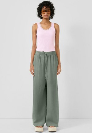 Frau mit lockigem Haar, die orangefarbene Sonnenbrille, ein pinkes ärmelloses Top, lockere grüne Hosen und Plateausandalen vor einem schlichten Hintergrund trägt.