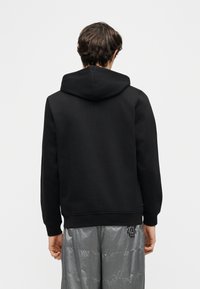 Άτομο που φοράει μαύρο φούτερ με κουκούλα και γκρι γυαλιστερό παντελόνι, στέκεται με την πλάτη προς το μέρος μας, μπροστά σε ένα απλό λευκό φόντο.