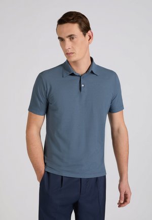 Uomo che indossa una polo a maniche corte blu e pantaloni blu scuro, in piedi con una mano in tasca, che guarda di lato su uno sfondo semplice.