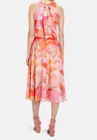 Kvinna i en ärmlös klänning med halterneck i rosa och orange blommönster, knuten i nacken, kombinerad med rosa pumps med ankelsnören, bakifrån.