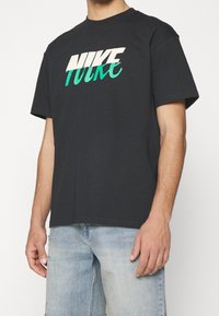Black cotton t-shirt with short sleeves, featuring "NIKE" logo in white and green cursive lettering across the chest. Classic fit design.