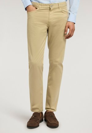 Uomo con pantaloni beige aderenti, camicia azzurro chiaro e mocassini di camoscio marroni, in piedi con una mano in tasca.