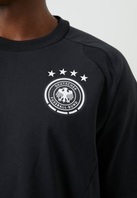 Maglia sportiva nera con un logo circolare bianco raffigurante un'aquila e cinque stelle sopra, con la scritta "Deutscher Fussball-Bund". Tessuto liscio.