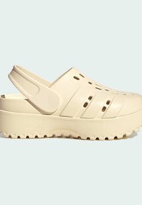Clogs en toile beige avec un bout fermé, des trous de ventilation circulaires et une sangle au dos. La semelle présente un motif texturé en crampons pour une meilleure adhérence.