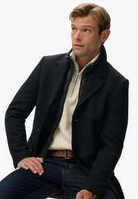 Abrigo de lana negro con un cuello estructurado sobre un suéter crema con cremallera en el cuello. Combinado con jeans oscuros y un cinturón marrón. Textura suave, ajuste entallado.