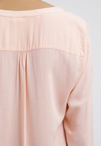 Blusa a maniche lunghe rosa pallido con dettaglio arricciato sul retro, realizzata in un tessuto leggero e morbido con una texture liscia.