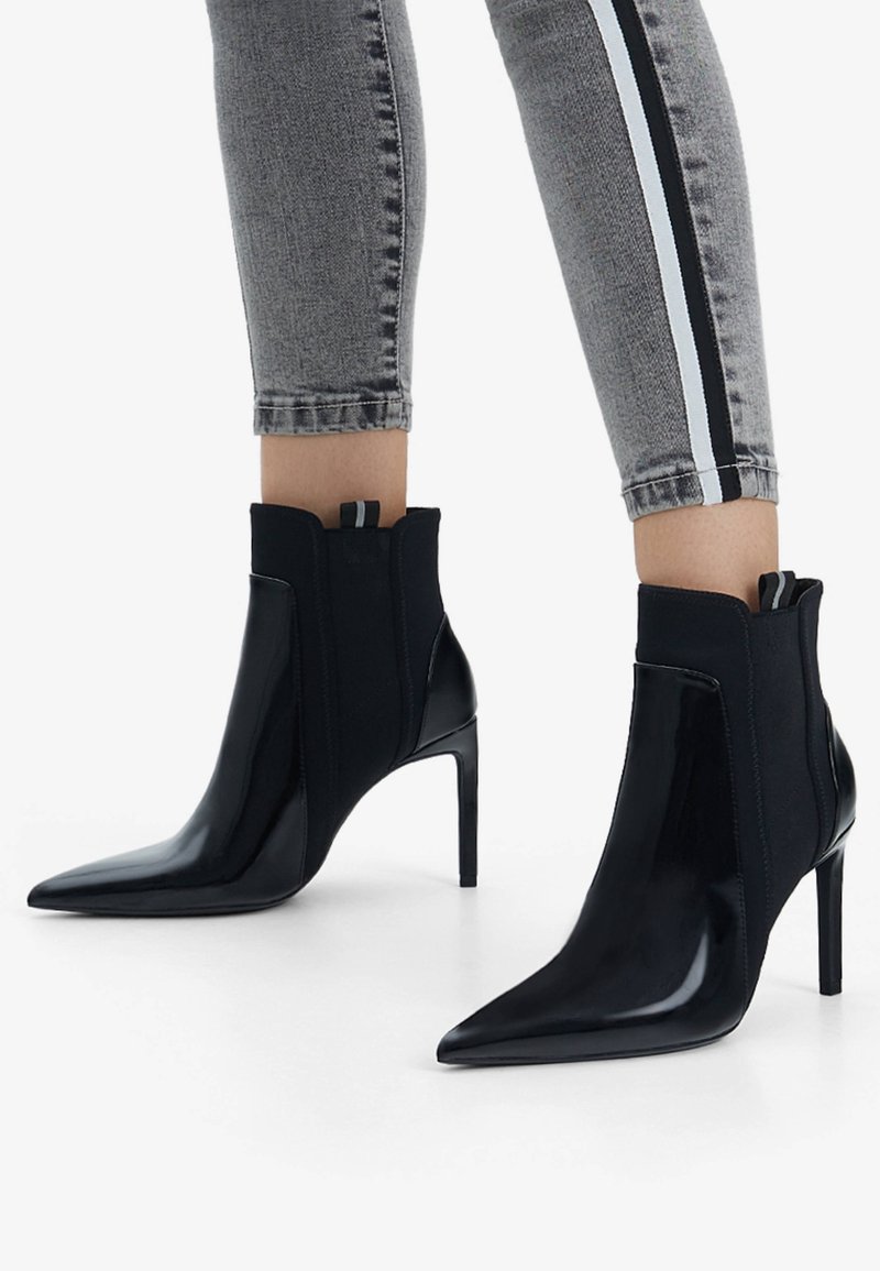 Jambes portant un jean skinny gris avec des bandes latérales et des bottines à talons hauts noires à bouts pointus avec des panneaux latéraux élastiques.