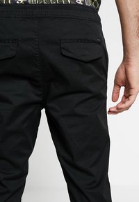 Man som bär svarta byxor med fickor med lock på baksidan, står mot en enkel ljus bakgrund, med höger hand avslappnat vid sidan.