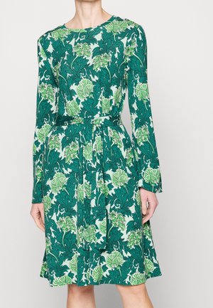 Sukienka o długości do kolan z długim rękawem, w zielony i jasnozielony wzór kwiatowy oraz liściasty, związaną paskiem w talii, noszona przez stojącą osobę.