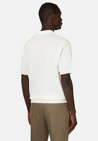 Homme à la peau foncée portant une chemise blanche unie à manches courtes et un pantalon marron, debout de dos devant un fond clair uni.