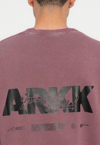 Übergröße weinroter Pullover mit großem schwarzem Aufdruck-Logo "ARKK" und Text auf dem Rücken. Gerippter Ausschnitt und strukturierter Stoff.