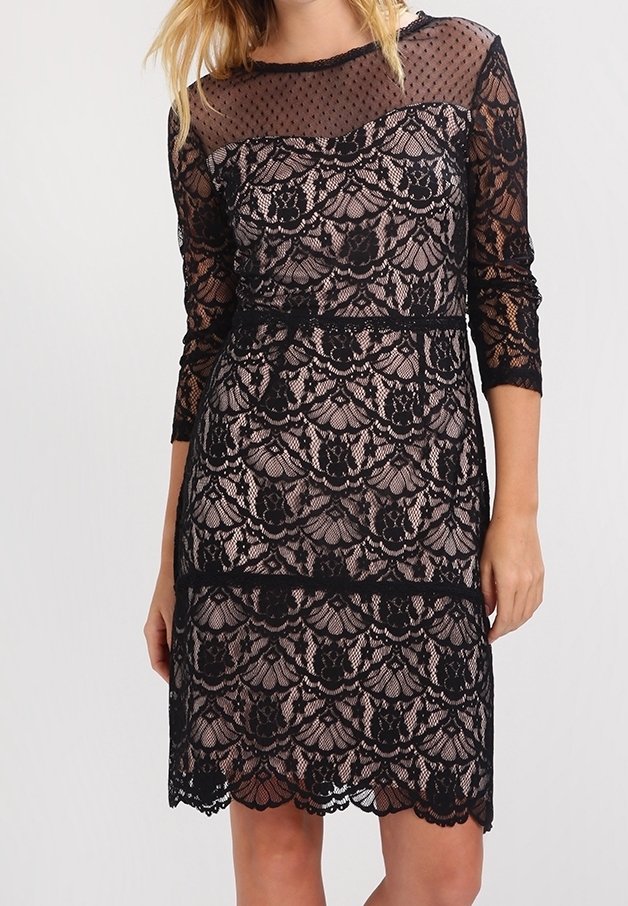 Femme portant une robe en dentelle noire avec une couche de base couleur chair, manches trois-quarts et un col transparent à pois.