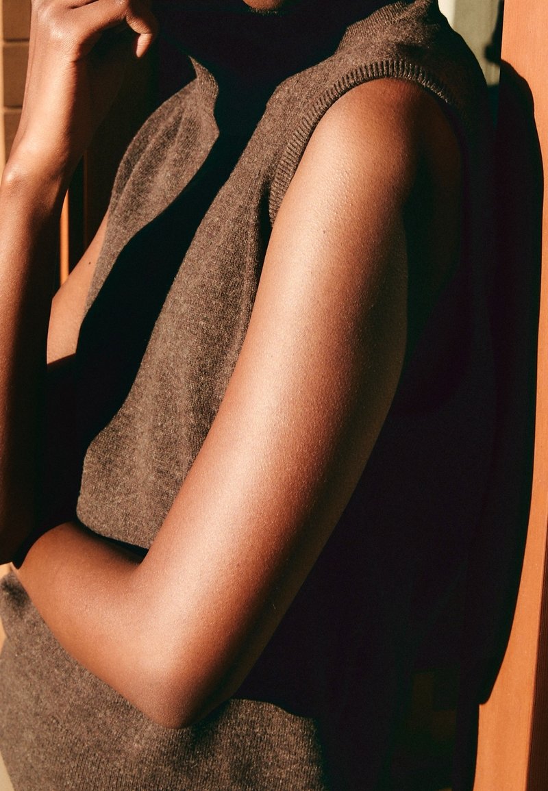 Personne portant un haut marron en tricot sans manches, reposant le bras sur l'abdomen avec la main près du visage, éclairage chaud projetant des ombres.