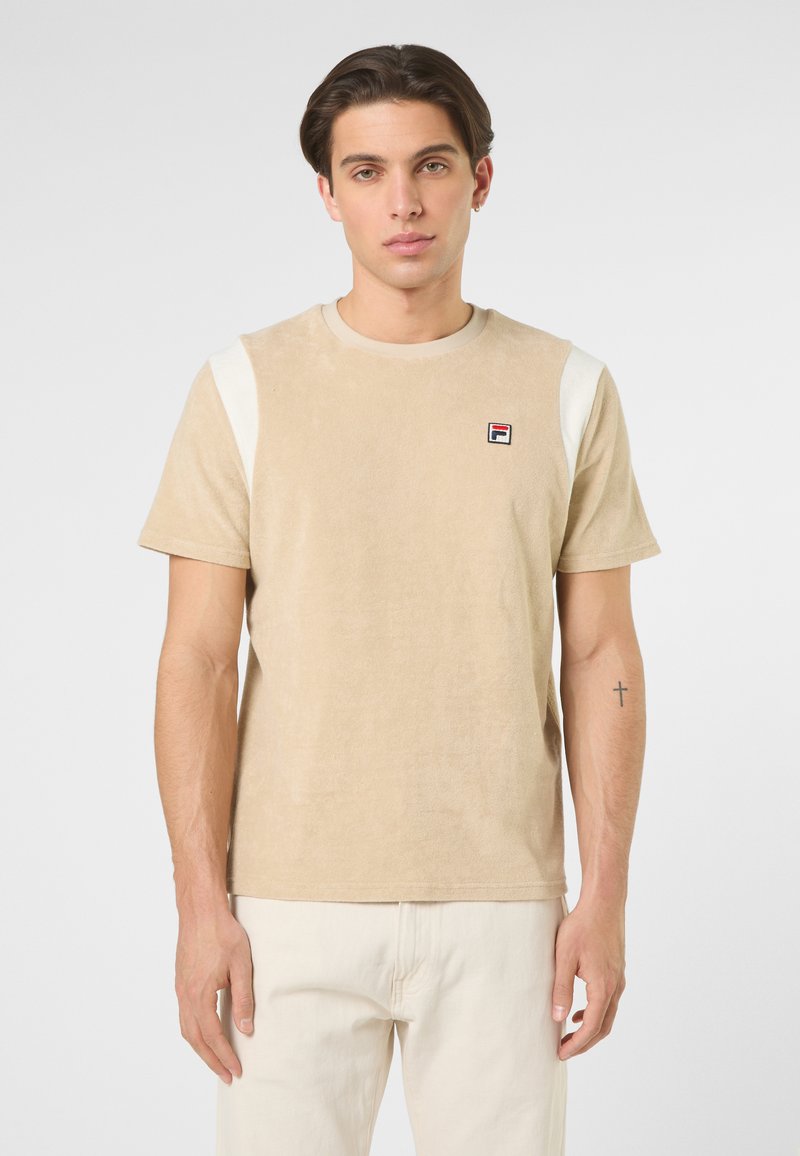 Jeune homme aux cheveux courts et foncés, portant un t-shirt beige à manches courtes avec des bandes blanches sur les épaules et un pantalon couleur crème, se tenant devant un fond uni.