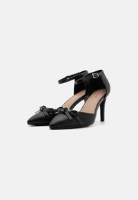 Black leather high-heeled shoes with pointed toe, ankle strap, and two decorative bows on the front. Smooth texture and sleek design.