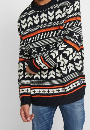 Man wearing black, white, and orange patterned knit sweater with geometric designs and blue jeans, arm bent across body.