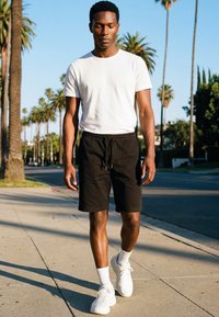 Man draagt een wit T-shirt, zwarte korte broek, witte sokken en sneakers, lopend op een zonnig trottoir omzoomd met palmbomen.