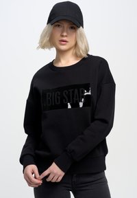 Černá mikina s lesklým černým obdélníkem, na kterém je nápis "BIG STAR". Model má na sobě odpovídající černou kšiltovku a tmavé džíny.