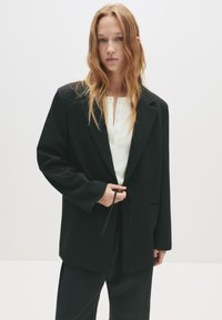 Jeune femme aux longs cheveux roux portant un blazer noir surdimensionné par-dessus un haut blanc à boutons, se tenant contre un fond clair uni.