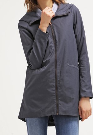 Femme portant une veste à capuche gris foncé zippée avec poches avant, associée à un jean bleu, sur un fond clair uni.