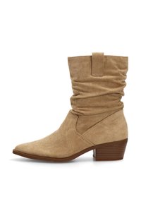 Bota de ante beige hasta la mitad de la pantorrilla con caña arrugada, tira de agarre, puntera afilada y tacón bajo apilado, sobre fondo blanco.
