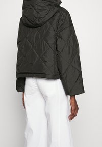 Giacca piumino trapuntata verde scuro con cappuccio, caratterizzata da una forma quadrata e maniche larghe, abbinata a pantaloni bianchi.