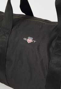 Svart duffelbag med teksturert stoff, glidelås og to korte håndtak. Har brodert logo med røde, hvite og blå detaljer.